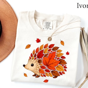 May include: An ivory t-shirt featuring a graphic of a hedgehog whose back is composed of various autumn-colored leaves in shades of red, orange, and brown. The shirt has a crew neckline and a Comfort Colors brand label printed on the inside collar.