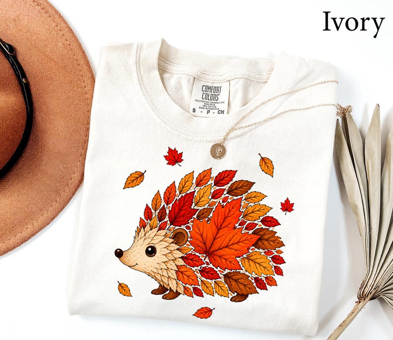 May include: An ivory t-shirt featuring a graphic of a hedgehog whose back is composed of various autumn-colored leaves in shades of red, orange, and brown. The shirt has a crew neckline and a Comfort Colors brand label printed on the inside collar.