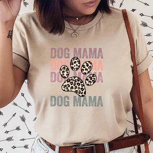 May include: A beige t-shirt with the text "DOG MAMA" printed in a layered, vintage style. A large paw print with a leopard print design is centered on the shirt.