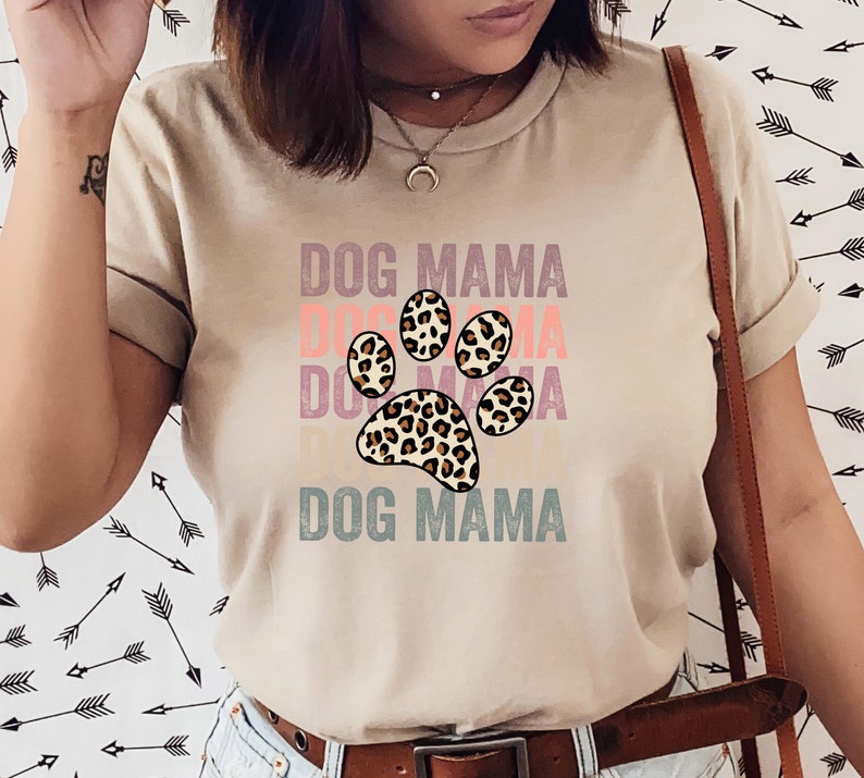 May include: A beige t-shirt with the text "DOG MAMA" printed in a layered, vintage style. A large paw print with a leopard print design is centered on the shirt.