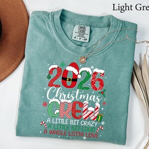 以下が含まれることがあります： 2026 Christmas Crew, A Little Bit Crazy, A Little Bit Loud, A Whole Lotta Loveというテキストがプリントされたライトグリーンの半袖Tシャツ。デザインには赤、緑、白のクリスマス柄、サンタの帽子、キャンディケインが含まれています。
