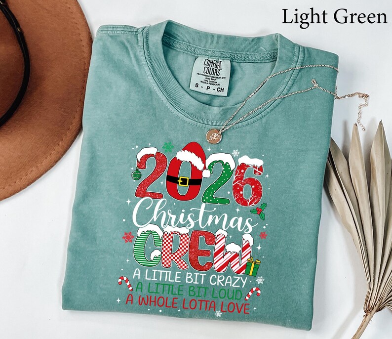 以下が含まれることがあります： 2026 Christmas Crew, A Little Bit Crazy, A Little Bit Loud, A Whole Lotta Loveというテキストがプリントされたライトグリーンの半袖Tシャツ。デザインには赤、緑、白のクリスマス柄、サンタの帽子、キャンディケインが含まれています。