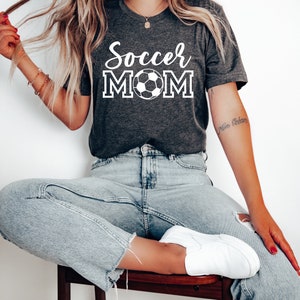 May include: A woman wearing a dark gray t-shirt with the text "Soccer Mom" and a white soccer ball graphic. She is wearing light blue jeans and white sneakers.
