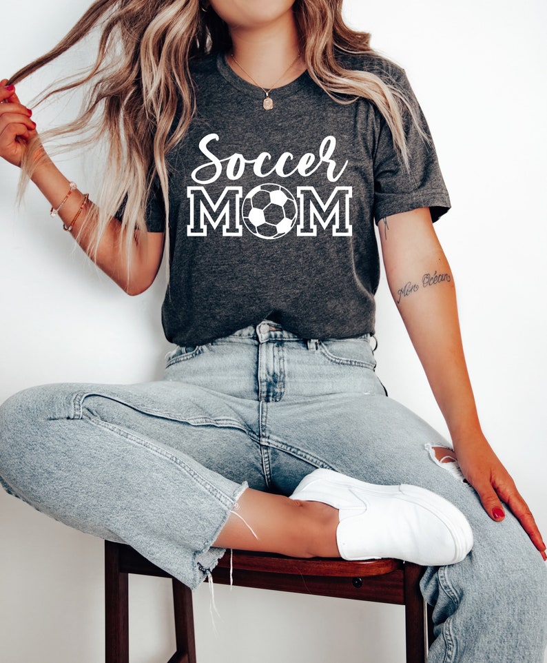 May include: A woman wearing a dark gray t-shirt with the text "Soccer Mom" and a white soccer ball graphic. She is wearing light blue jeans and white sneakers.