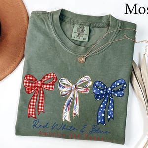 May include: A moss green short-sleeve t-shirt featuring three decorative bows printed across the chest. The left bow is red and white gingham, the center is white with red and blue accents, and the right is blue with white stars. Text reads Red White & Blue.