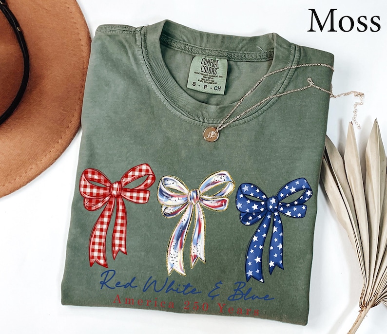 May include: A moss green short-sleeve t-shirt featuring three decorative bows printed across the chest. The left bow is red and white gingham, the center is white with red and blue accents, and the right is blue with white stars. Text reads Red White & Blue.