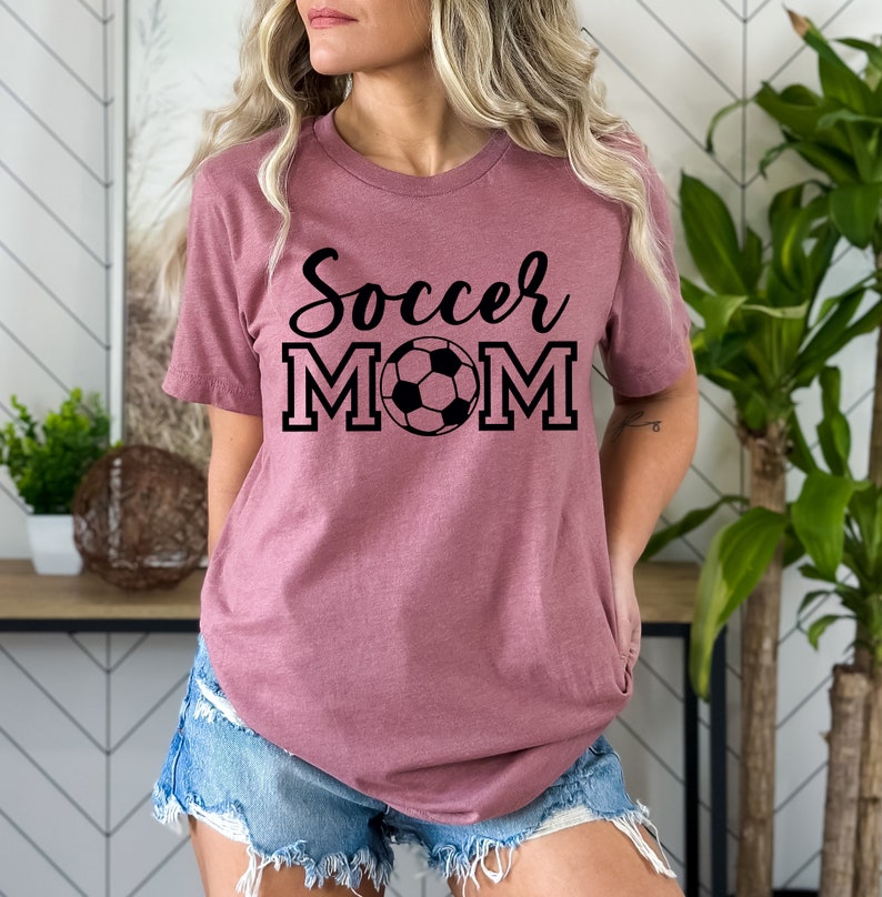 May include: A mauve t-shirt with the text "Soccer Mom" in black lettering with a black and white soccer ball in the middle of the word "Mom".