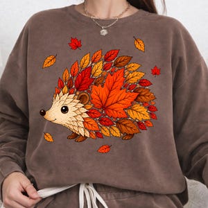 May include: A brown crewneck sweatshirt featuring a graphic illustration of a hedgehog. The hedgehog's body is composed of various autumn-colored leaves in shades of red, orange, and yellow. Several loose leaves are scattered around the hedgehog.