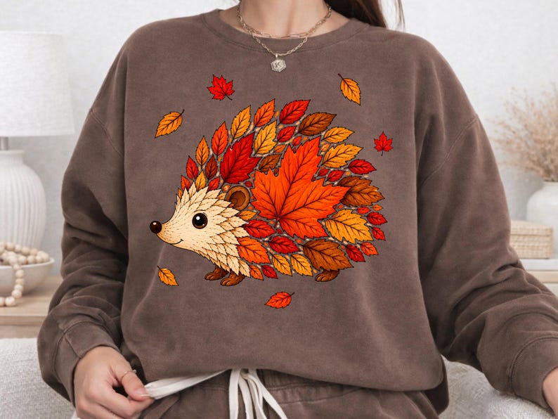 May include: A brown crewneck sweatshirt featuring a graphic illustration of a hedgehog. The hedgehog's body is composed of various autumn-colored leaves in shades of red, orange, and yellow. Several loose leaves are scattered around the hedgehog.