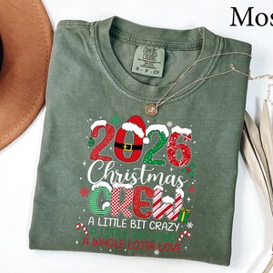 以下が含まれることがあります： モスグリーンの半袖Tシャツ。2026 Christmas Crew, A Little Bit Crazy, A Little Bit Loud, A Whole Lotta Loveというテキストがプリントされています。赤と緑の模様、サンタの帽子、小さなクリスマスのアイコンがデザインに含まれています。