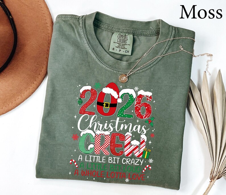 以下が含まれることがあります： モスグリーンの半袖Tシャツ。2026 Christmas Crew, A Little Bit Crazy, A Little Bit Loud, A Whole Lotta Loveというテキストがプリントされています。赤と緑の模様、サンタの帽子、小さなクリスマスのアイコンがデザインに含まれています。