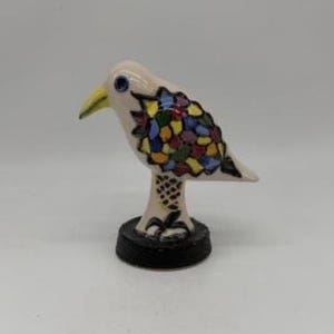 Puede incluir: Figura de pájaro de cerámica con cuerpo color crema, pico amarillo y base negra. Las alas del pájaro están decoradas con patrones coloridos tipo mosaico. La figura es un objeto decorativo.