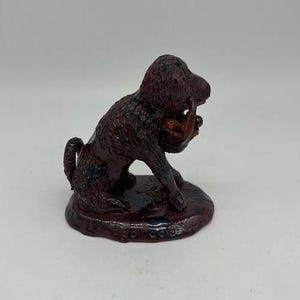 May include: A dark brown figurine of a dog-like creature with a textured surface. The sculpture is sitting on a round base. The dog is holding a smaller figure in its mouth.
