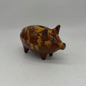 May include: A ceramic piggy bank in a brown and yellow speckled pattern. The pig has a slot on its back for inserting coins and a small, upturned tail. The piggy bank is a classic design for saving money.