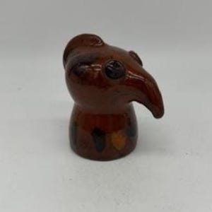 May include: A brown ceramic figurine shaped like a plague doctor's mask. The figurine has a long, curved beak, small eye holes, and a rounded head. The base of the figurine is cylindrical with dark spots.