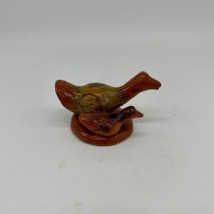 May include: A handcrafted ceramic duck figurine. The sculpture features a larger duck atop a smaller duck, both with orange and brown tones. The ducks are perched on a circular base, creating a charming decorative piece.