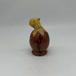May include: A ceramic figurine of a bird with a yellow head and a brown body. The bird has a detailed, textured design on its chest and is perched on a small base. The figurine is painted with a glossy finish.