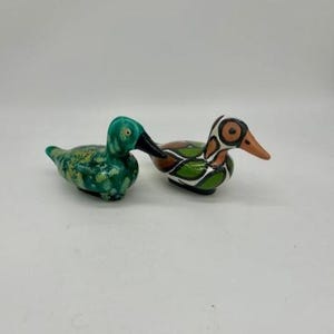 Puede incluir: Dos figuras de patos de cerámica. Una es verde con detalles amarillos, la otra tiene un patrón marrón, verde y blanco. Ambos tienen picos negros y están sobre un fondo blanco. Estos patos decorativos son para la decoración del hogar.