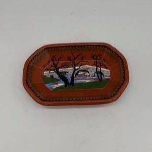 Puede incluir: Una pequeña bandeja decorativa rectangular con un fondo marrón rojizo. La bandeja presenta una escena de paisaje pintada con árboles, un puente y agua. La bandeja tiene un borde festoneado.