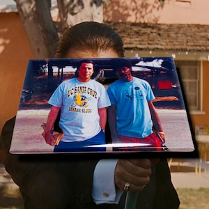 May include: A close-up of a photo of two people standing outside. The person on the left is wearing a white t-shirt with the text "UC Santa Cruz Banana Slugs". The person on the right is wearing a blue t-shirt. The photo is framed in a black frame.