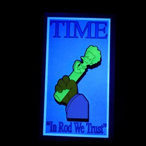 May include: A green and blue enamel pin featuring a cartoon character holding a microphone. The pin is shaped like a magazine cover with the word "TIME" printed in blue at the top and the text "In Rod We Trust" printed in blue at the bottom.