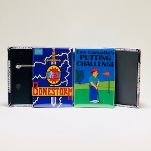 May include: Four rectangular pinback buttons. One is black, and the others feature colorful graphics. One button has the text "BONESTORM" with a sword and fist graphic. Another says "Lee Carvallo's Putting Challenge" with a cartoon golfer.