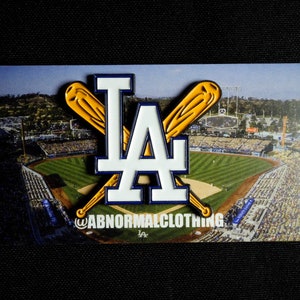 May include: A white enamel pin with a blue outline of the letters "LA" with two crossed yellow baseball bats behind the letters. The pin is on a background of a baseball stadium with a crowd of people in the stands.