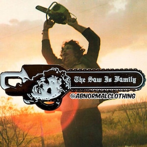 May include: A black and white enamel pin shaped like a chainsaw with the text "The Saw Is Family" and the social media handle "@ABNORMALCLOTHING".