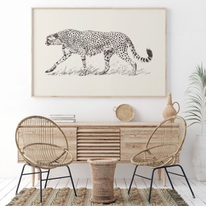 May include: A black and white line drawing of a cheetah walking through tall grass. The drawing is framed and hanging on a white wall above a wooden cabinet with two wicker chairs and a small wicker stool.