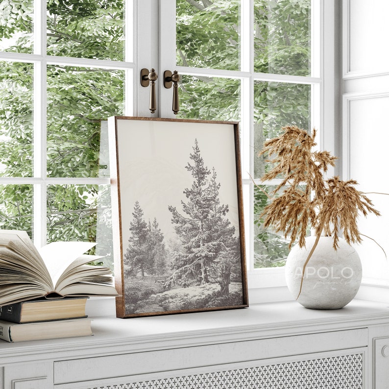 Op de afbeelding: Een zwart-wit ingelijste print van een besneeuwde bos sc&egrave;ne met hoge dennenbomen. De print hangt aan een witte muur boven een witte commode met een decoratieve vaas en gedroogde bloemen.