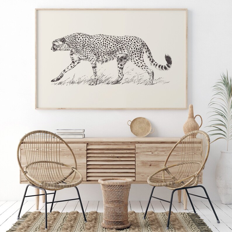 May include: A black and white line drawing of a cheetah walking through tall grass. The drawing is framed and hanging on a white wall above a wooden cabinet with two wicker chairs and a small wicker stool.