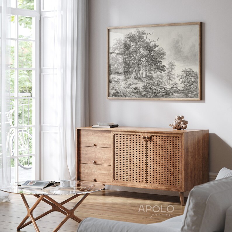 May include: A wooden cabinet with three drawers and two doors. The doors are made of woven rattan and have wooden knobs. The cabinet is sitting on a wooden floor in a living room with a white wall and a window with white curtains. A black and white framed print of a forest scene hangs on the wall above the cabinet. A glass coffee table with a wooden frame sits in front of the cabinet.