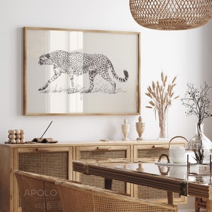 May include: A black and white illustration of a cheetah walking through tall grass, framed in a light wood frame. The frame is hanging on a white wall above a wooden cabinet with woven rattan doors.