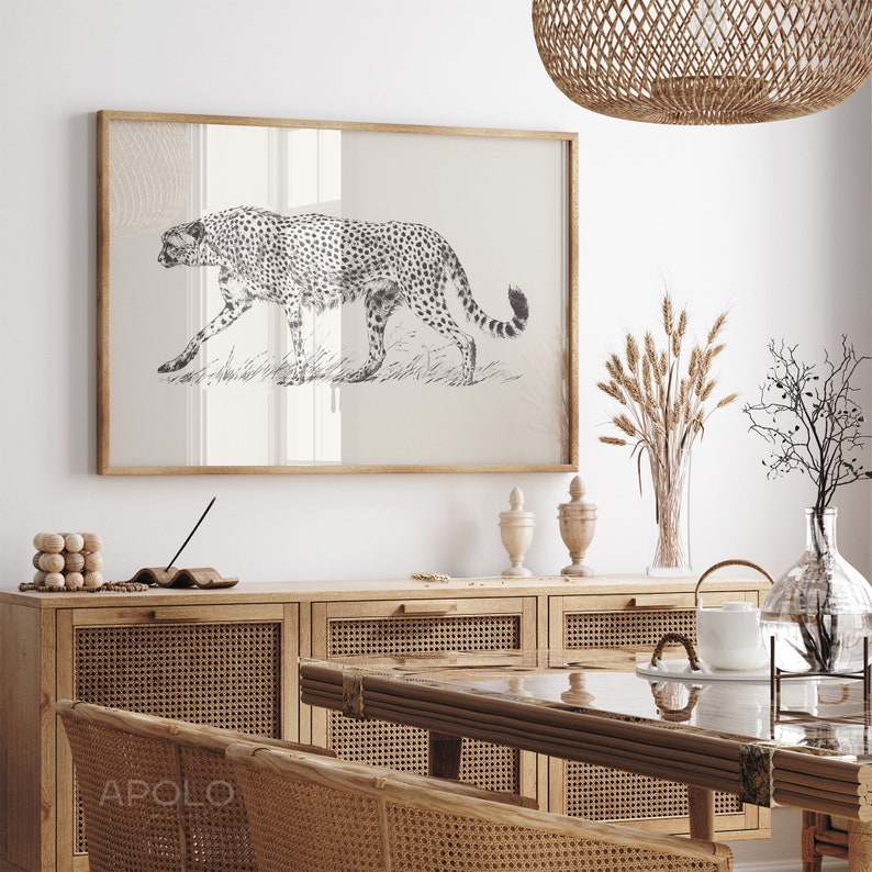 May include: A black and white illustration of a cheetah walking through tall grass, framed in a light wood frame. The frame is hanging on a white wall above a wooden cabinet with woven rattan doors.