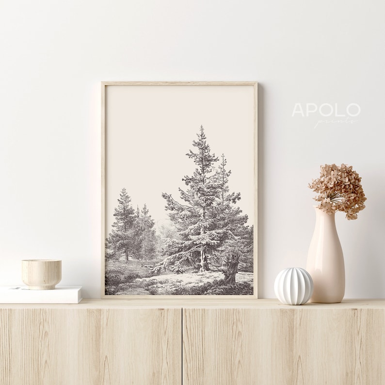Op de afbeelding: Een ingelijste zwart-witprint van een besneeuwde bos sc&egrave;ne met drie dennenbomen. De print is op een licht beige achtergrond en is ingelijst in een licht houten lijst.