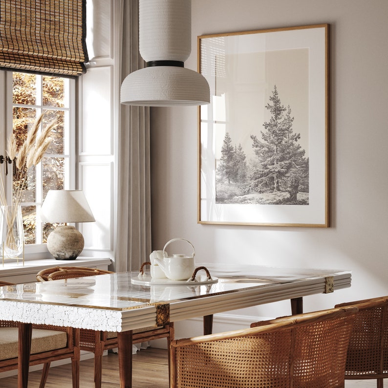 Op de afbeelding: Een witte keramische hanglamp hangt boven een eettafel met een glazen blad en een wit houten frame. De tafel is gedekt met twee stoelen met gevlochten rieten zittingen en rugleuningen. Een ingelijste print van een bos sc&egrave;ne hangt aan de muur achter de tafel.