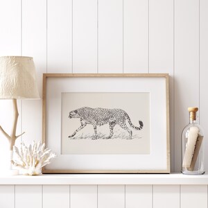 May include: A framed black and white illustration of a cheetah walking through tall grass. The frame is made of light wood and has a white mat.