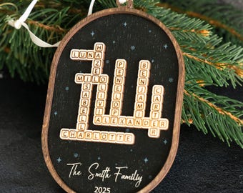 May include: Wooden family ornament with a black background and a Scrabble-style design. The ornament features names like Luna, Milo, and Charlotte, with "The Smith Family 2025" written below. It has a white ribbon for hanging.