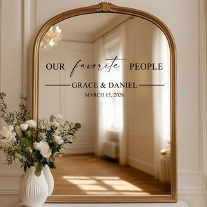 May include: A gold-framed arched mirror with the text "OUR favorite PEOPLE - GRACE & DANIEL - MARCH 15, 2026". A white vase with white flowers and greenery is in front of the mirror.