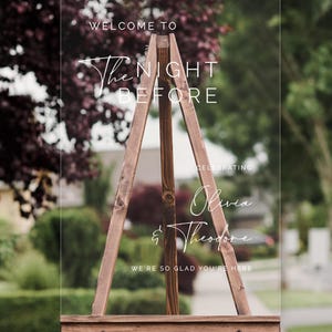 Pu&ograve; includere: Un cartello di benvenuto in acrilico trasparente con il testo "Welcome to The Night Before Celebrating Olivia & Theodore We're so glad you're here" su un cavalletto di legno.