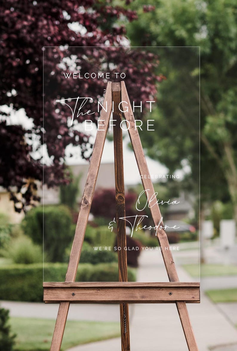Pu&ograve; includere: Un cartello di benvenuto in acrilico trasparente con il testo "Welcome to The Night Before Celebrating Olivia & Theodore We're so glad you're here" su un cavalletto di legno.