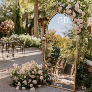 May include: A large, arched mirror with a gold frame, decorated with pink roses and greenery. The mirror reflects an outdoor setting with tables and chairs. Text on the mirror reads "We're so glad you're here" and names with a date.