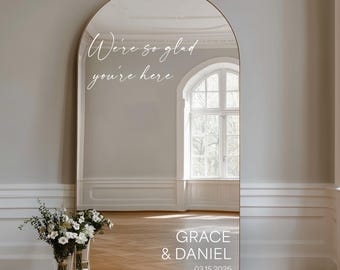 May include: An arched, gold-framed mirror with the text "We're so glad you're here" in elegant script. The mirror reflects a room with a window and a bouquet of flowers. The names "GRACE & DANIEL" and the date 03.15.2026 are at the bottom.