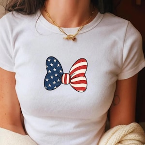 May include: White t-shirt featuring a patriotic bow tie design. The bow tie is split into two sections: one side with a blue background and white stars, and the other with red and white stripes. The bow tie is centered on the shirt.