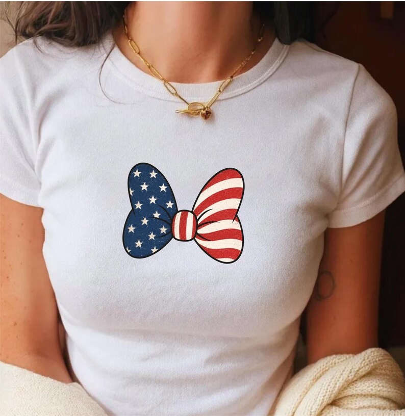 May include: White t-shirt featuring a patriotic bow tie design. The bow tie is split into two sections: one side with a blue background and white stars, and the other with red and white stripes. The bow tie is centered on the shirt.