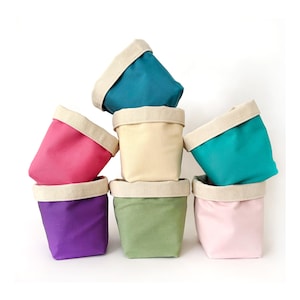 以下が含まれることがあります： 7つの小さなカラフルな布製収納容器のスタック。各容器は、ティール、ピンク、紫、緑、クリームなど、異なる色で、自然色の折り返しリムが付いています。容器は視覚的に魅力的な積み重ね構成で配置されています。