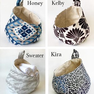 Puede incluir: Cuatro cestas de almacenamiento de tela colgantes con diferentes patrones. Las cestas est&aacute;n etiquetadas como "Honey", "Kelby", "Sweater" y "Kira".