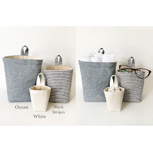 May include: Three hanging fabric storage baskets in ocean blue, black and white stripes, and white. The ocean blue basket holds rolled towels. The black and white striped basket holds eyeglasses. The white basket is small and holds cotton swabs.