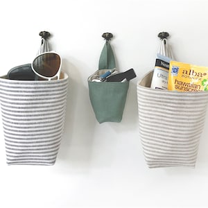 May include: Three hanging fabric storage baskets. Two are gray and white striped, holding sunglasses and sunscreen. The third is solid green, containing keys and a small flashlight. All are suspended from decorative metal hooks.