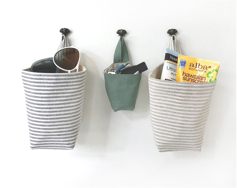 May include: Three hanging fabric storage baskets. Two are gray and white striped, holding sunglasses and sunscreen. The third is solid green, containing keys and a small flashlight. All are suspended from decorative metal hooks.
